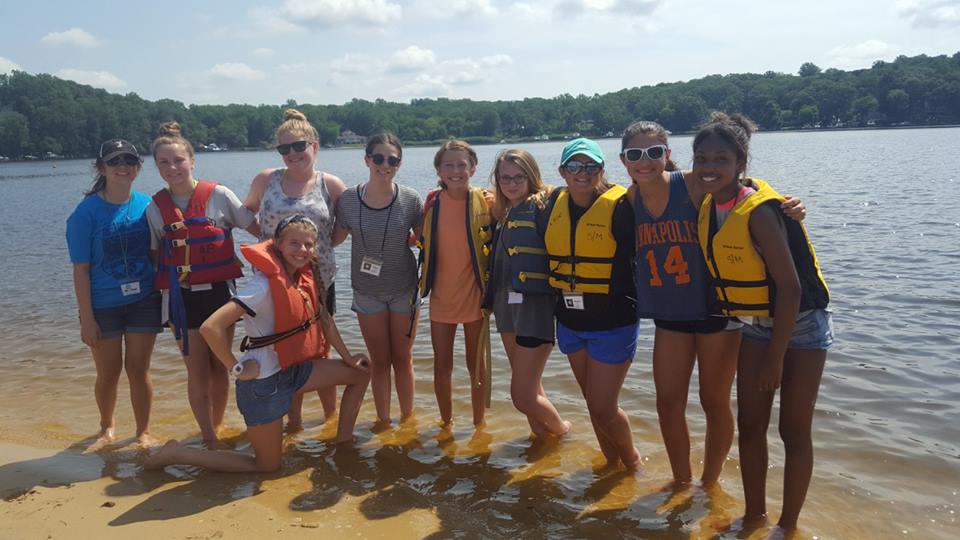 10 campers standing on sand at the edge of a river. 